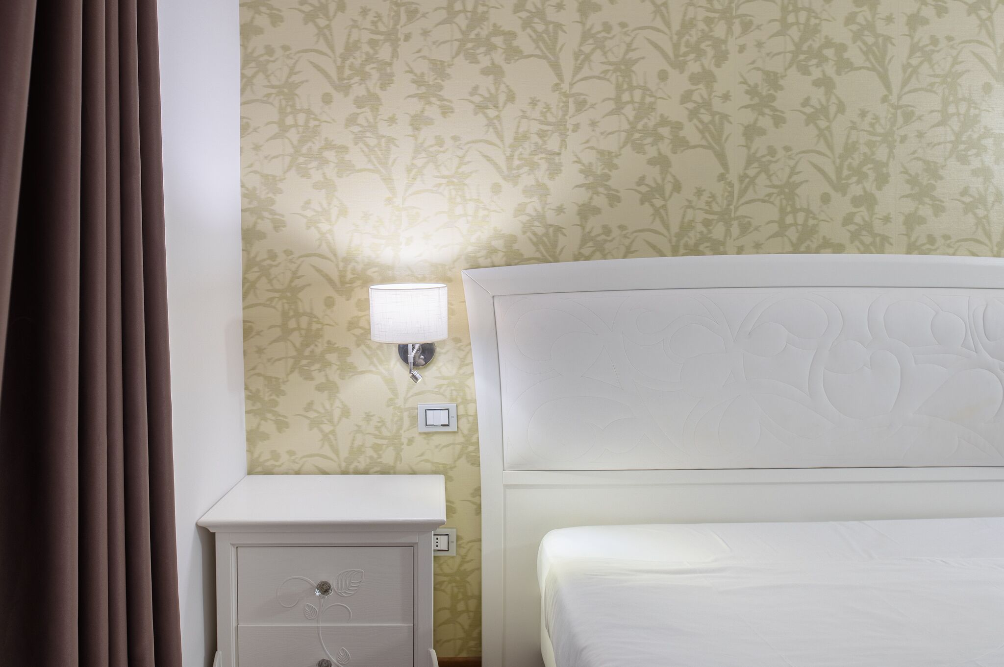 A hotel room with a bed, nightstand, lamp, and floral wallpaper.