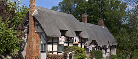A traditional half-timbered house with a thatched roof, brick chimneys, and a garden with various plants and flowers.