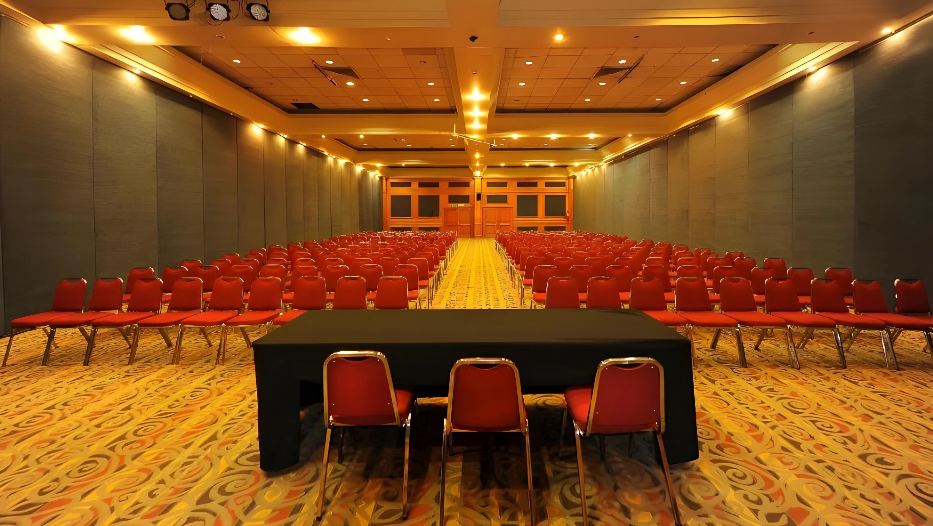Un salón de conferencias con filas de sillas rojas, una mesa grande al frente y alfombra estampada.