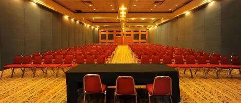 Un salón de conferencias con filas de sillas rojas, una mesa grande al frente y alfombra estampada.