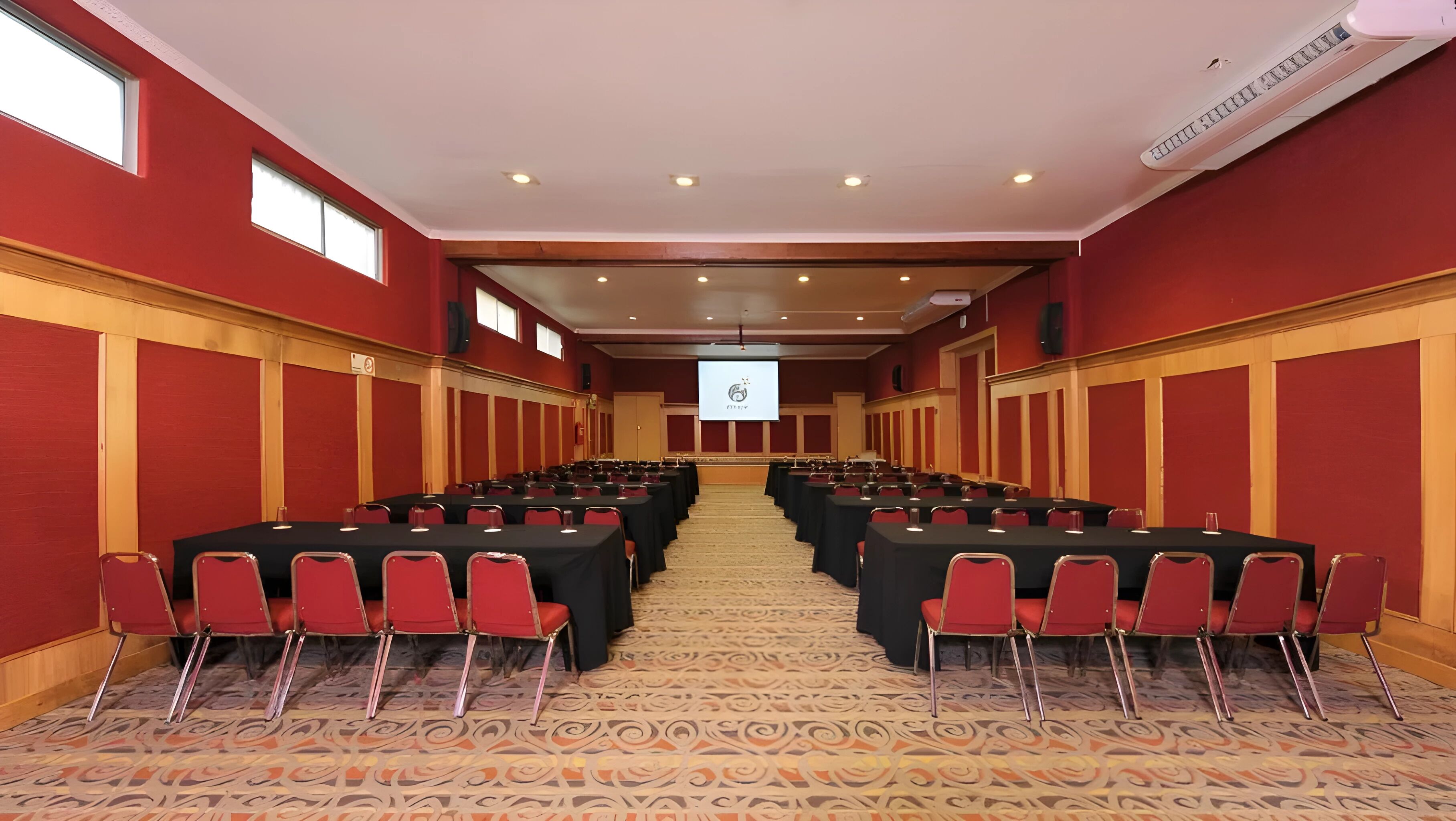 Un salón de conferencias con filas de mesas y sillas, suelo alfombrado y una pantalla de proyector al frente.