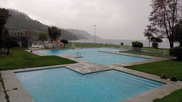 A rectangular pool with a surrounding deck, a small play area, and a view of the sea and hills in the background.