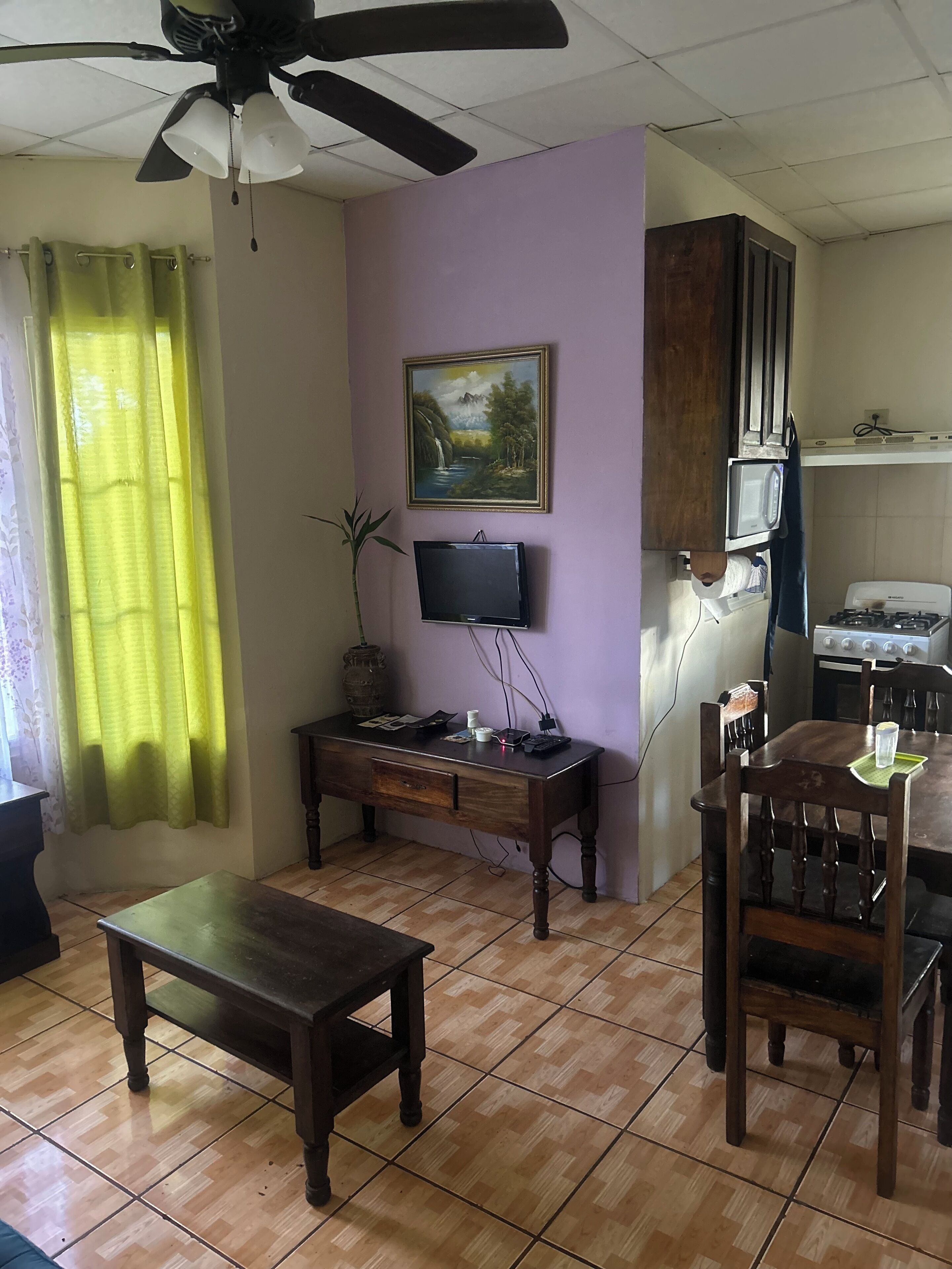A living room with a wooden table, a TV on a stand, a ceiling fan, and a kitchen area with a stove and microwave.