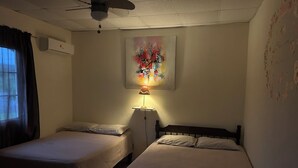 Un dormitorio con dos camas individuales, ventilador de techo, unidad de aire acondicionado y un cuadro en la pared.