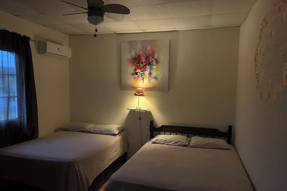 Un dormitorio con dos camas individuales, ventilador de techo, unidad de aire acondicionado y un cuadro en la pared.