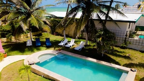 Ein rechteckiger Swimmingpool mit klarem, blauem Wasser, umgeben von einer Rasenfläche mit Palmen und einigen Liegestühlen.