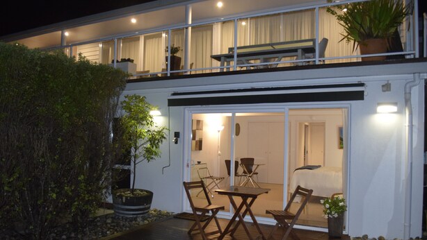 Ein modernes, zweistöckiges Haus mit Holzterrasse, Sitzgelegenheit im Freien und einer großen Pflanzung.