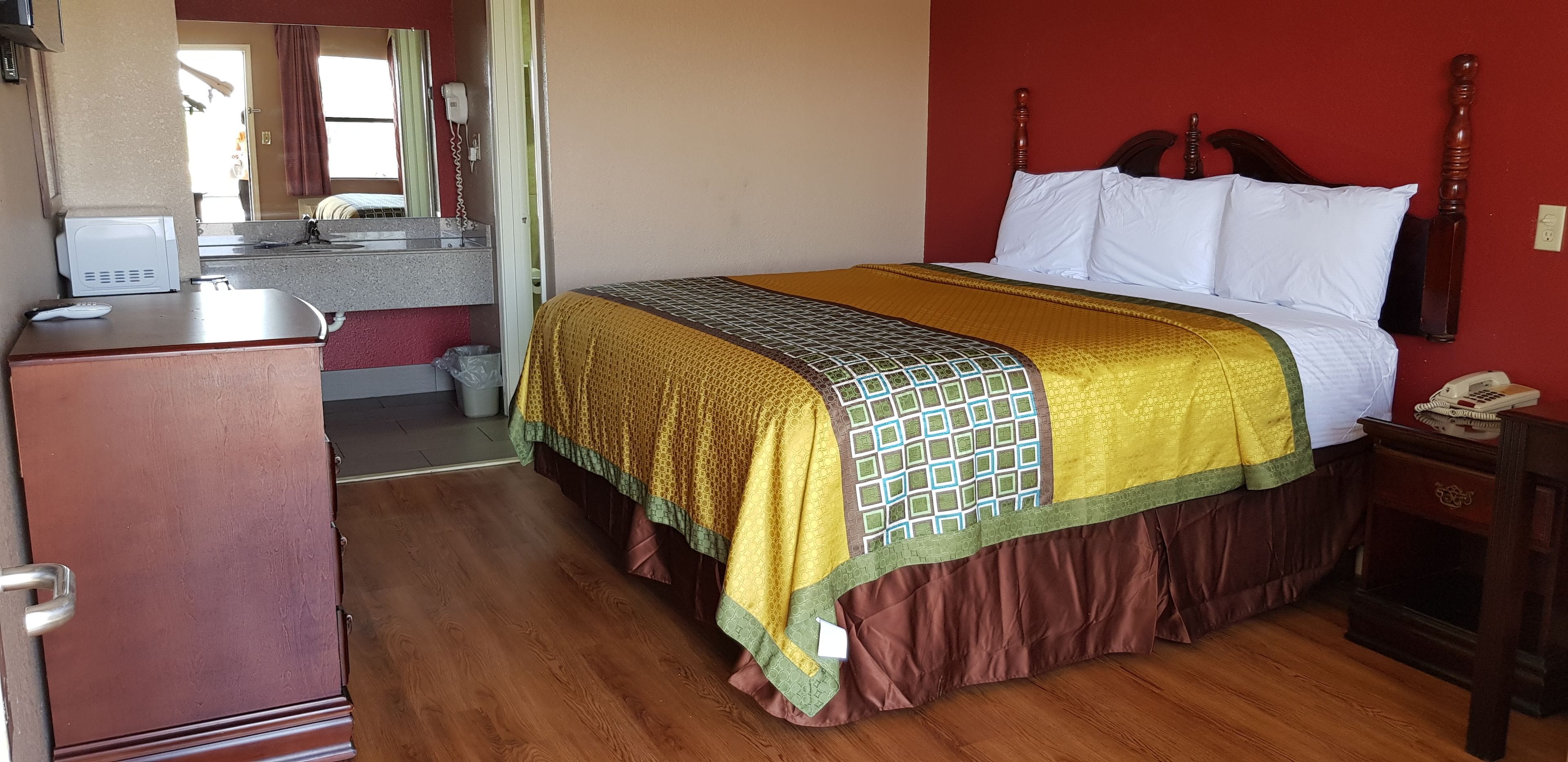 Ein Hotelzimmer mit einem hölzernen Bett, einem Nachttisch mit Telefon, einem kleinen Schreibtisch mit Computer und einem Spiegel.