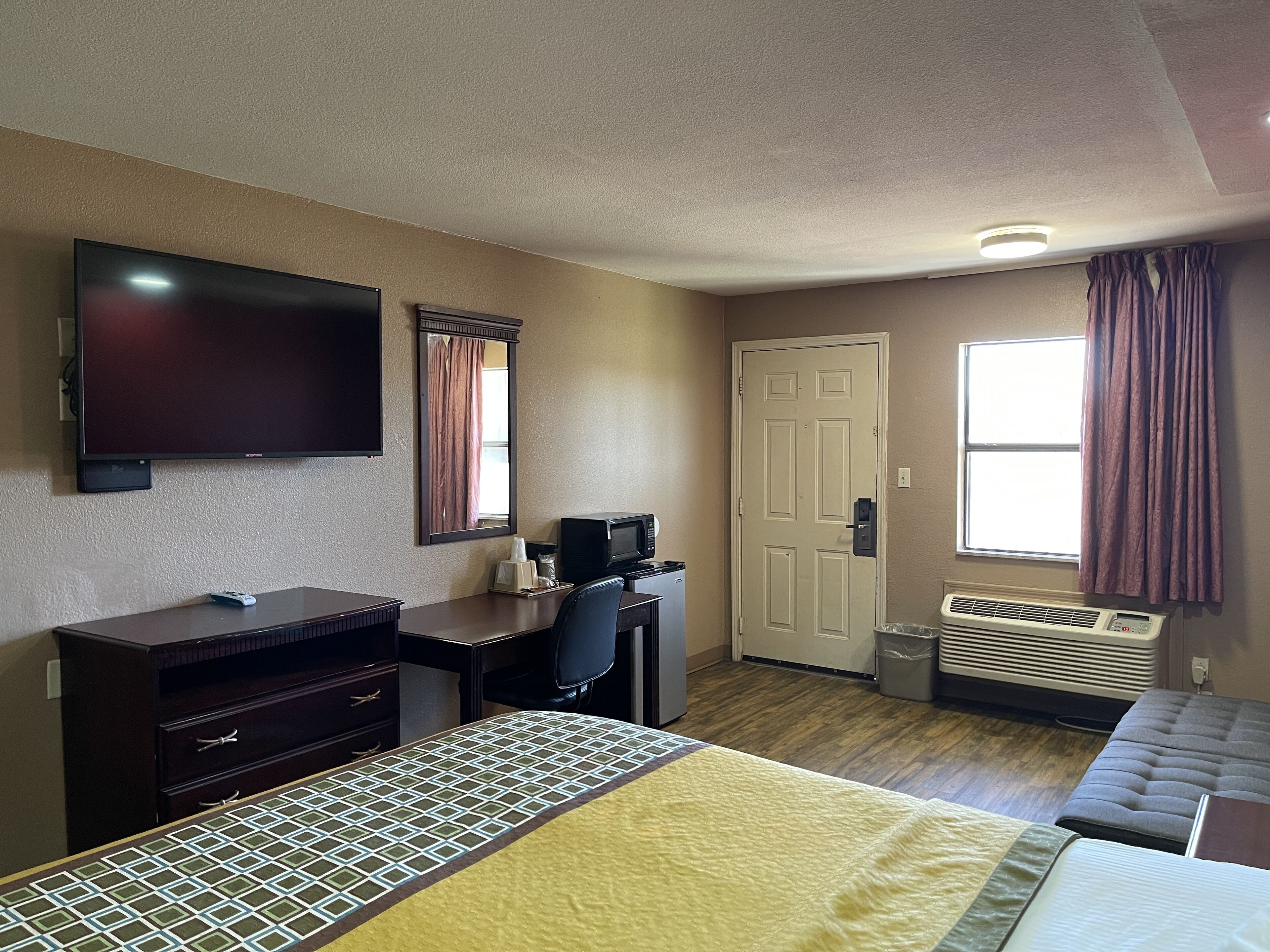 A hotel room with a bed, a desk with a computer, a television, a door, and an air conditioning unit.