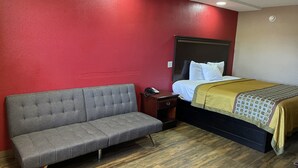 A hotel room with a red wall, a grey sofa, a wooden floor, a bed with white bedding, and a nightstand with a phone. - Deluxe Inn (West Memphis)