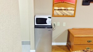 A compact room with a refrigerator, a wooden dresser, and a mirror on the wall. - Deluxe Inn (West Memphis)