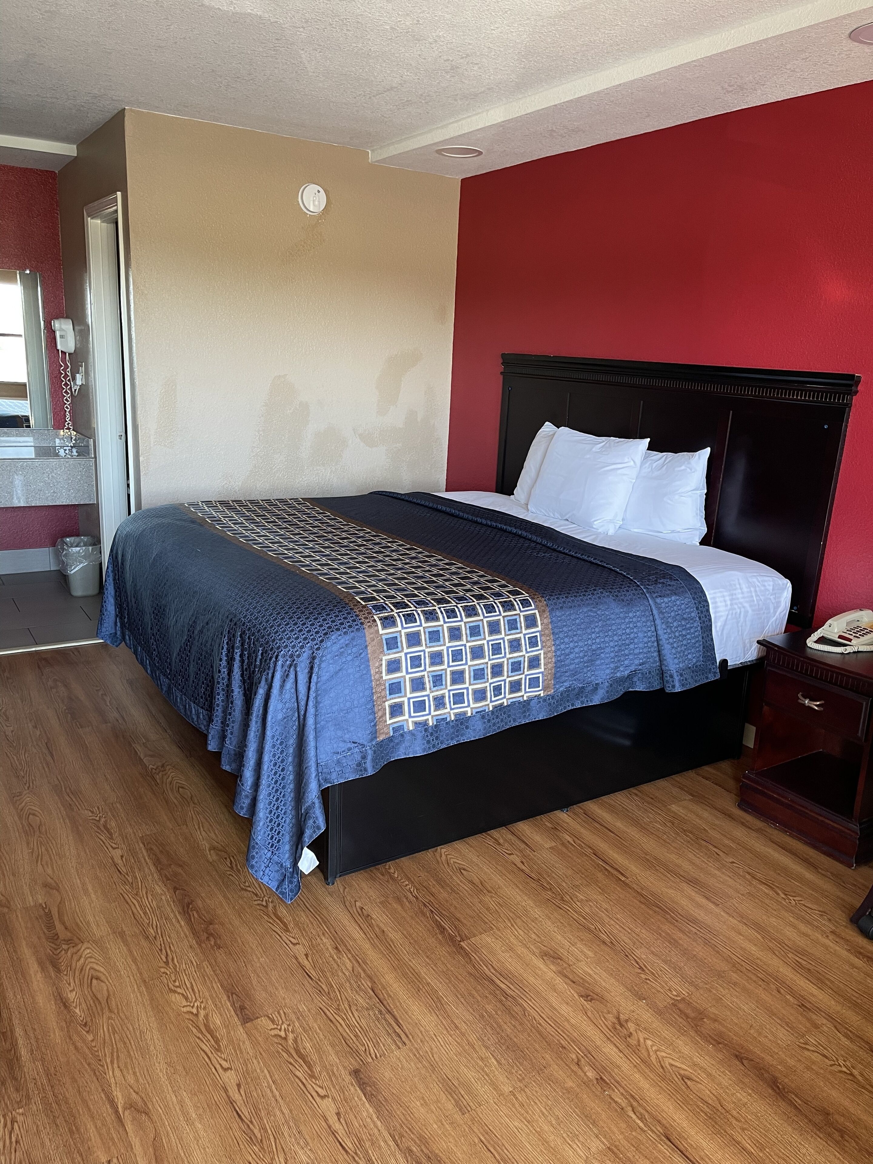 A hotel room with a bed, wooden flooring, a red wall, and a beige wall.