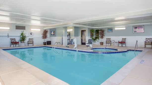 An indoor swimming pool with clear blue water, surrounded by beige tiled flooring and walls. There are several chairs and potted plants around the pool area.