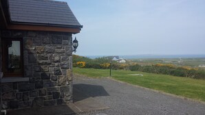 A stone building with a dark roof, a window with curtains, and a view of the sea and distant houses.