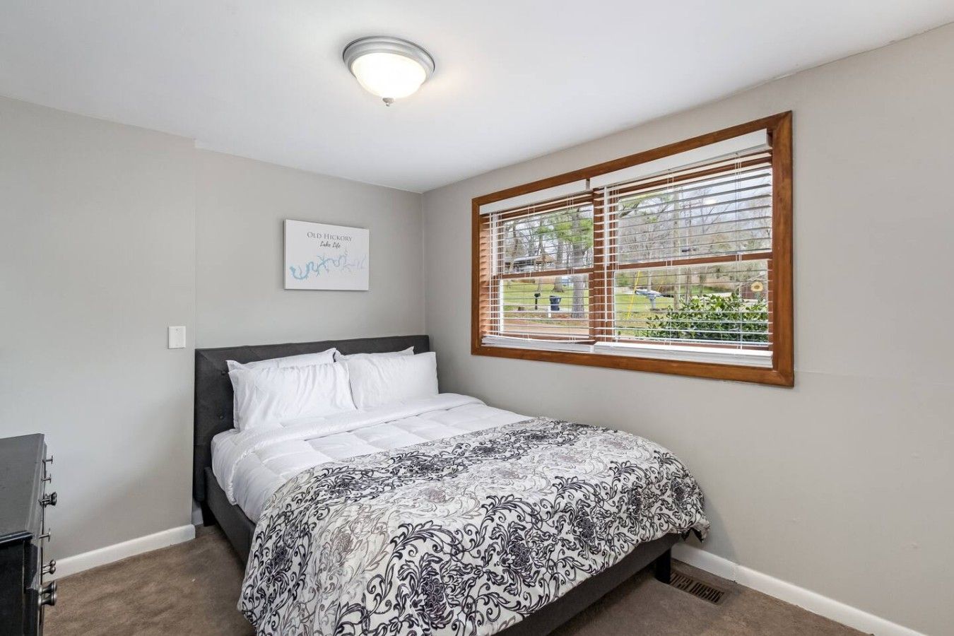 Une chambre à coucher avec un lit, une fenêtre avec des stores, un luminaire au plafond et un panneau fixé au mur.
