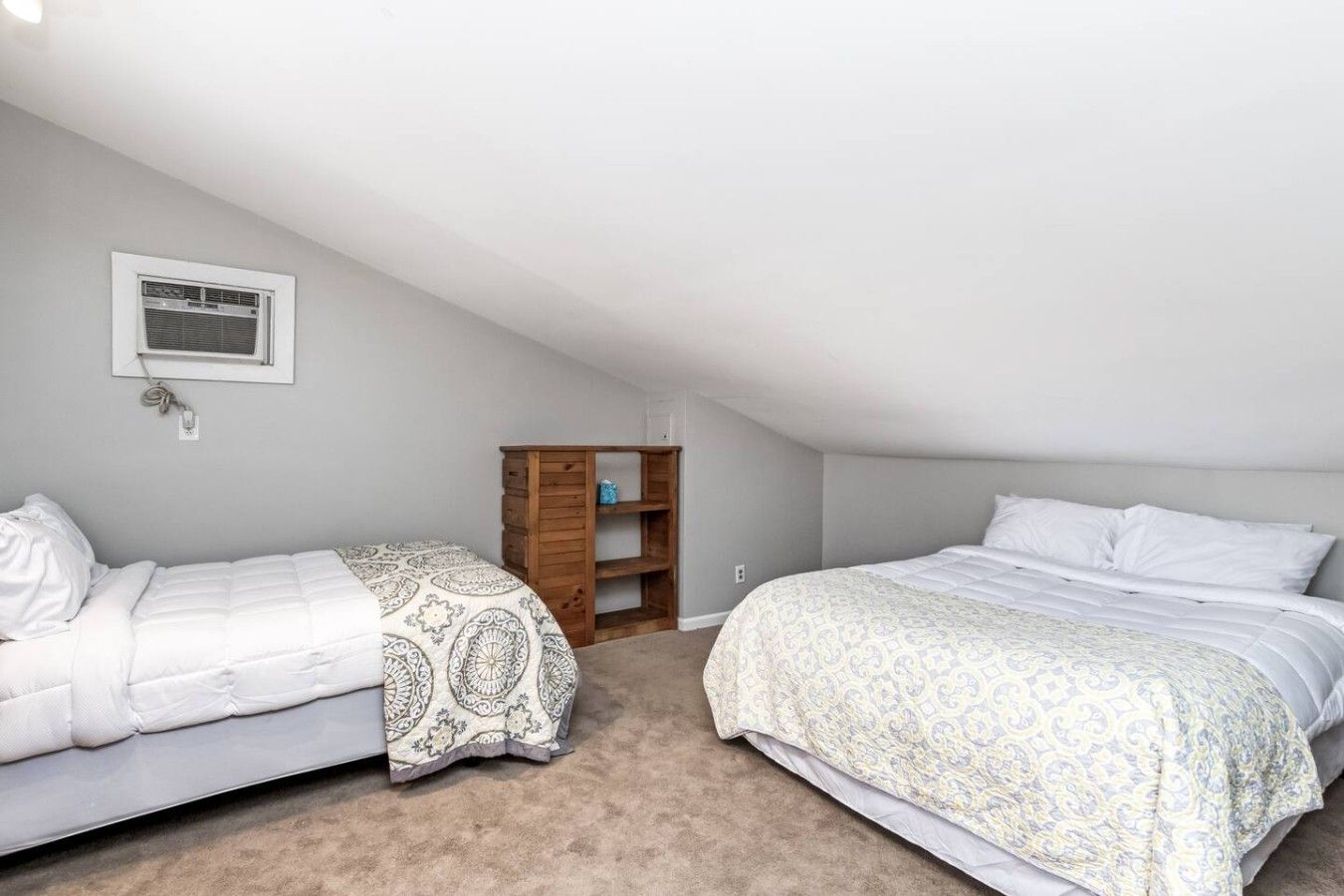 Une chambre avec deux lits, une étagère en bois et une fenêtre équipée d’un appareil de climatisation.