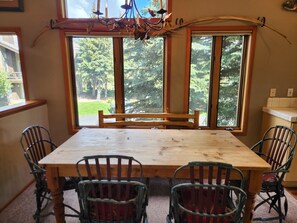 Un comedor con mesa y sillas de madera, ventanas amplias con vista a árboles y una araña.
