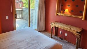 Ein Schlafzimmer mit einem Bett, einem Holznachttisch, einem Spiegel mit Fischmotiven und Blick auf ein rotes Gebäude durch eine Glastür.