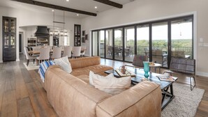 A spacious living room with a large leather sofa, a dining area with chairs, a kitchen with cabinets, and large sliding glass doors leading to a balcony with a view. - High-end Home on 33 Acres to Roam, Short Drive to Downtown. (Austin)