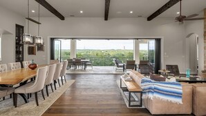 A spacious dining area with a wooden table and chairs, a sofa with a patterned throw, and a balcony with a view of the greenery. - High-end Home on 33 Acres to Roam, Short Drive to Downtown. (Austin)