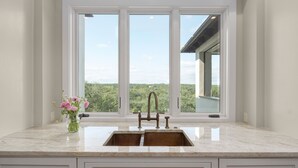 A kitchen with a double sink, a vase with flowers, and a view of greenery through large windows. - High-end Home on 33 Acres to Roam, Short Drive to Downtown. (Austin)