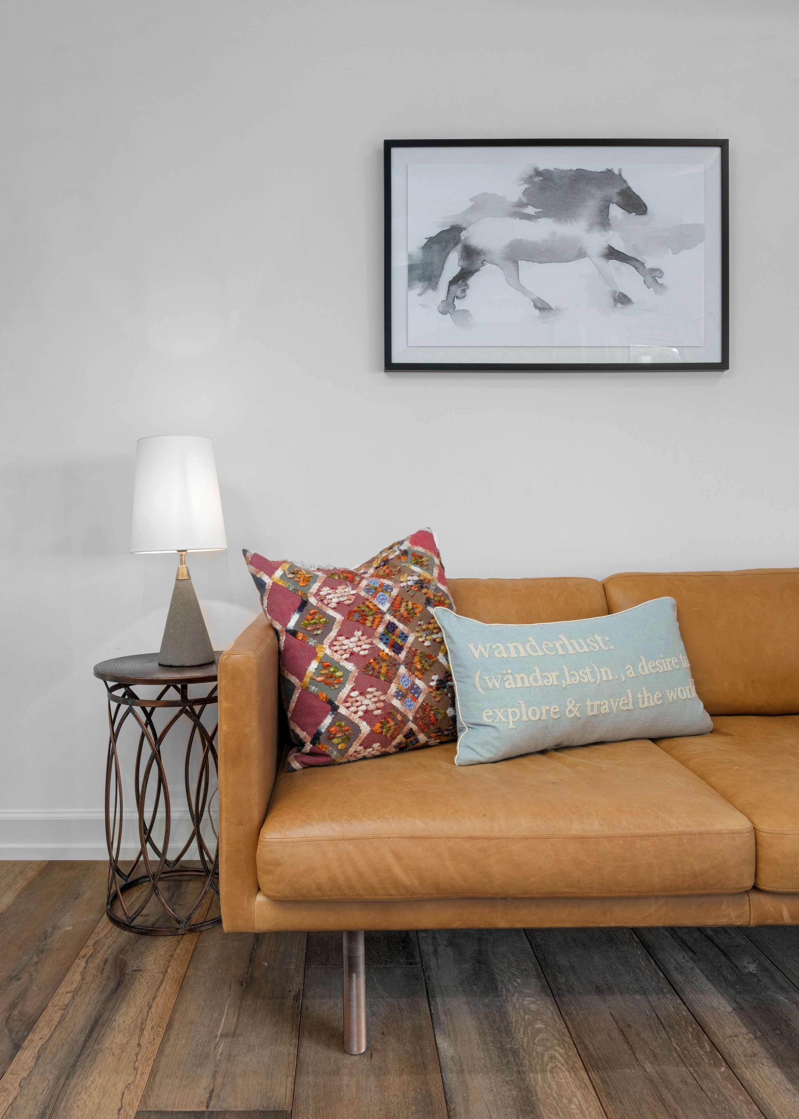 Ein braunes Ledersofa mit zwei Kissen, eines mit Patchwork-Muster und das andere mit Schrift. Darüber an der Wand hängt ein Schwarz-Weiß-Bild von einem Pferd in einem Rahmen.