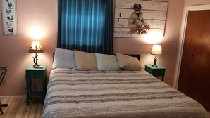 Een netjes opgemaakt bed met een gestreept dekbed, twee nachtkastjes met lampen, een blauw gordijn en een muur met een schilderij van een haan.