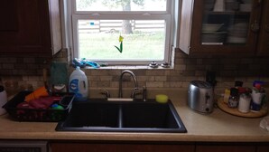 Een keuken met een spoelbak, een raam met uitzicht op een tuin, en verschillende keukenbenodigdheden op het aanrecht.