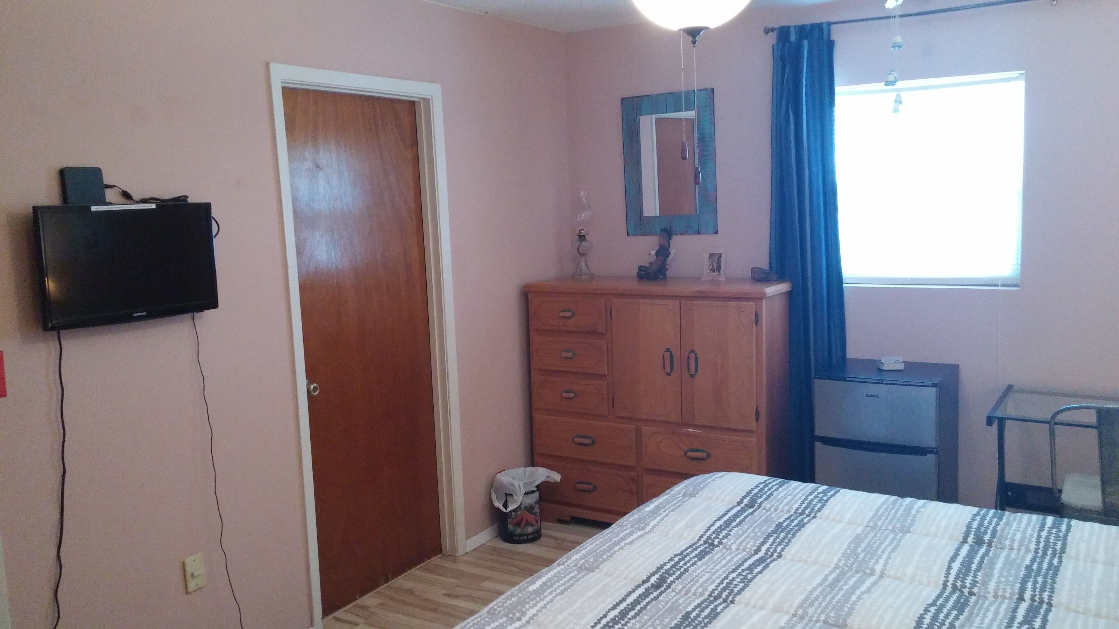 A bedroom with a bed, a wooden dresser, a television mounted on the wall, and a window with curtains.