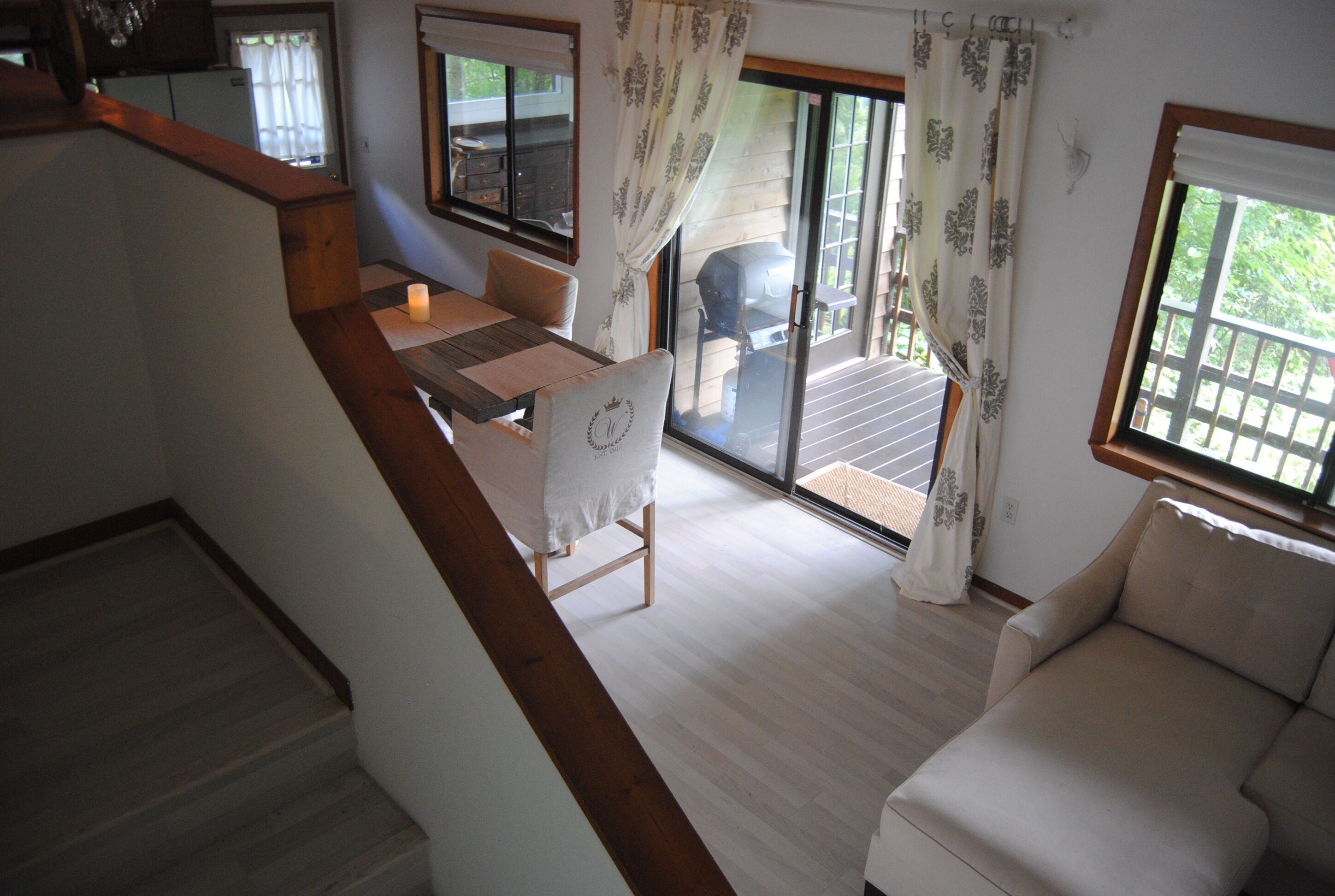 A spacious living area with a wooden staircase, a dining table with chairs, a sofa, and a sliding glass door leading to an outdoor deck.
