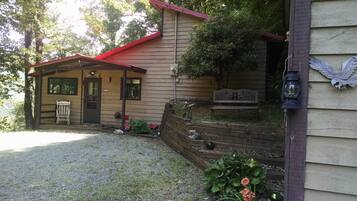 A rustic cabin with a red roof, a porch with a bench, and a mailbox.