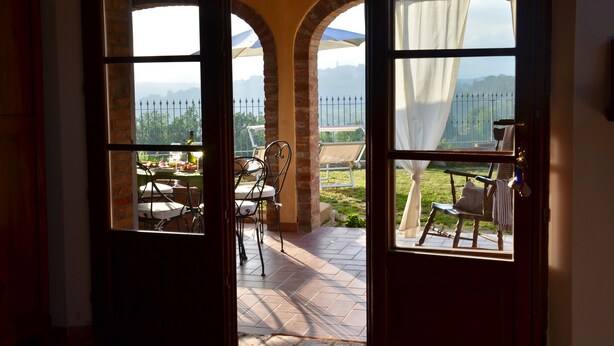 Eine geöffnete Tür führt zu einer Terrasse mit Tisch, Stühlen und einer Sitzbank, mit Blick auf einen Zaun und viel Grün.
