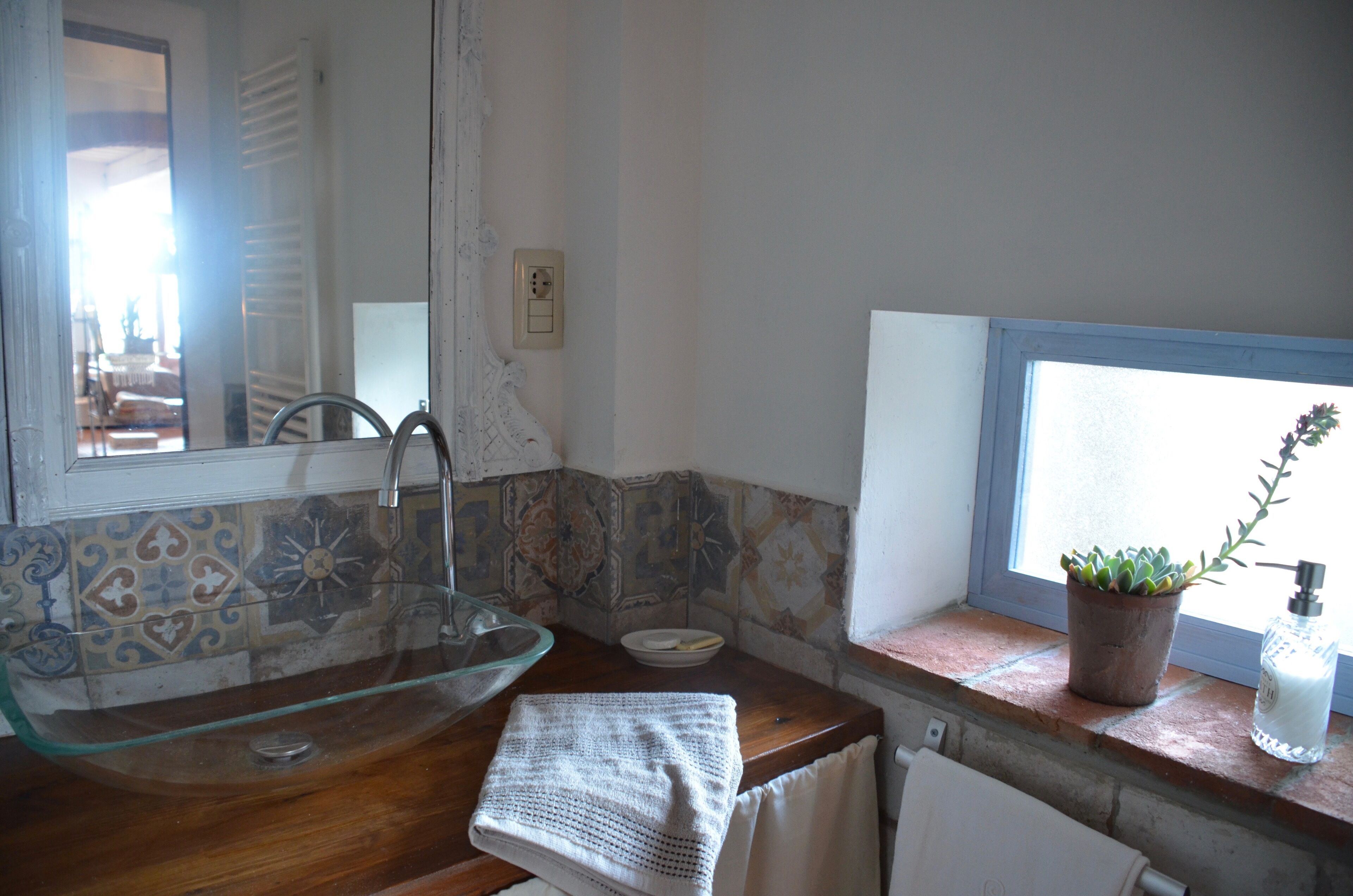Ein Badezimmer mit einem Glaswaschbecken, einem Handtuch, einer Topfpflanze und einem Fenster.