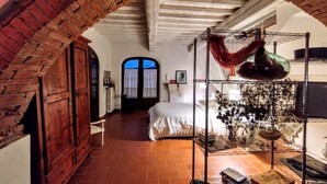 Ein Zimmer mit Backsteinbogen, einem Bett mit weißer Bettwäsche, einem Metallregal mit diversen Gegenständen und einem Fenster mit Vorhängen.