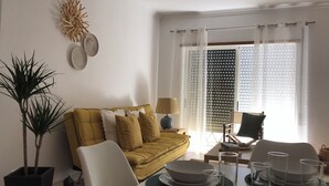 A living room with a yellow sofa, a dining table set with plates and glasses, a lamp, and a large window with blinds. - Casa Yeti - Beach House (Armação de Pêra)