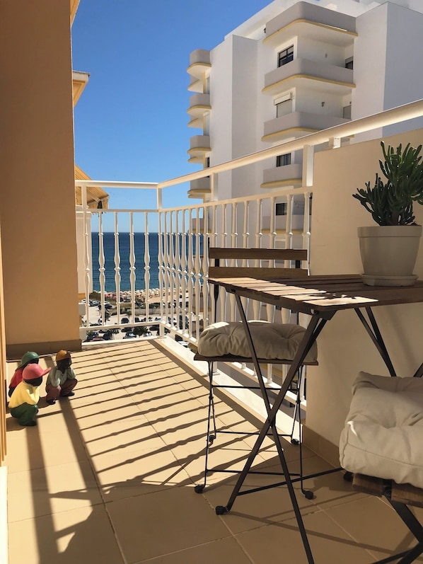 A balcony with a view of the sea, a white railing, a table with a cushion, and a potted plant.