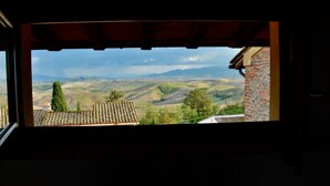 Une vue depuis une fenêtre donnant sur un paysage vallonné, avec un grand arbre et un mur de briques.