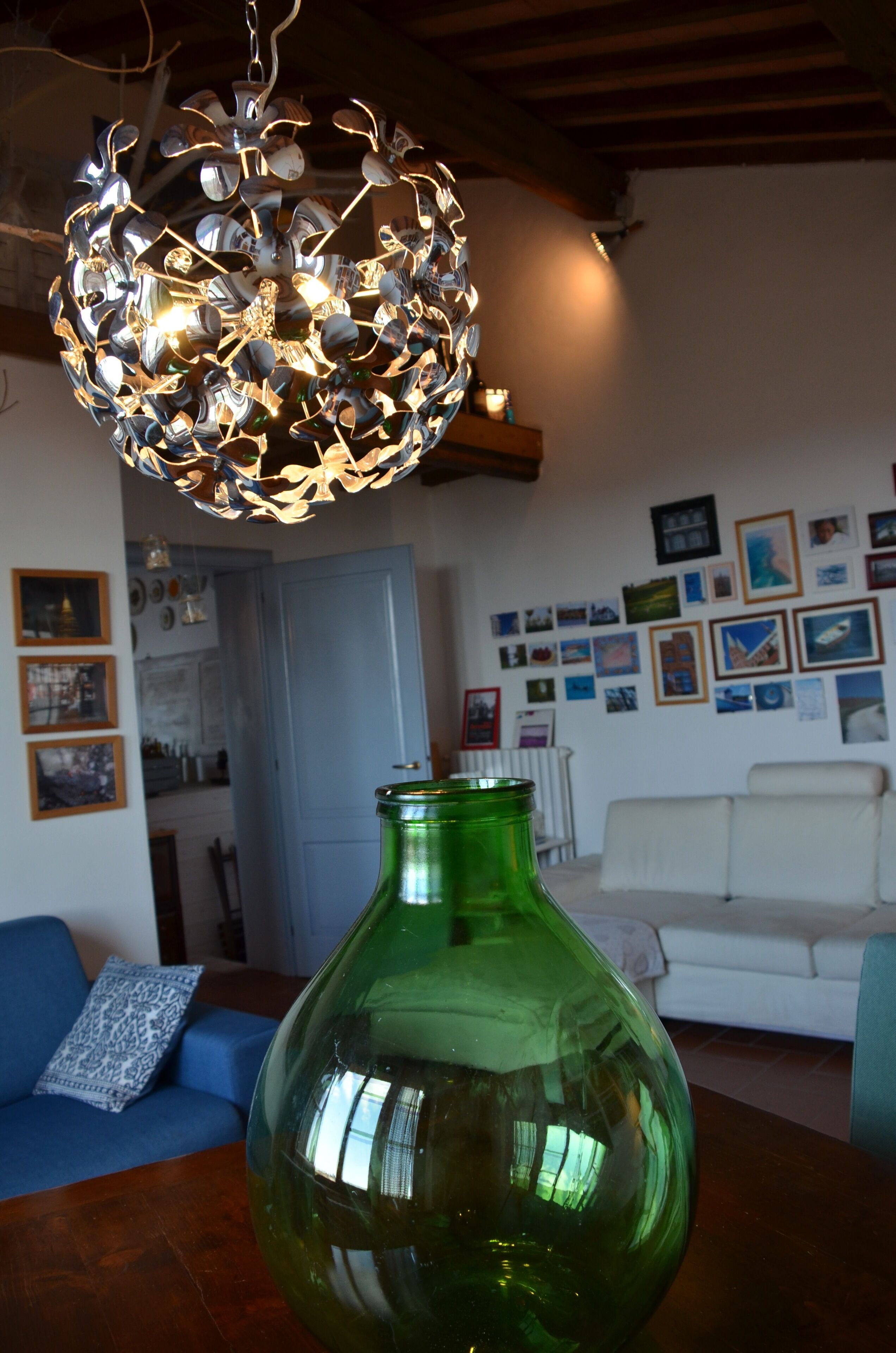 Ein Wohnzimmer mit einer großen grünen Vase auf einem Holztisch, einem blauen Sofa mit gemusterten Kissen, einem weißen Sofa und einer Wand, die mit gerahmten Bildern geschmückt ist.
