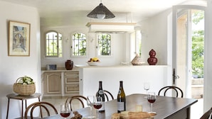 A dining area with a wooden table, chairs, wine glasses, and a bottle of wine. There are windows with views of greenery outside. - Luxury Provencal villa with heated pool and gym in St Paul (Saint-Paul-De-Vence)
