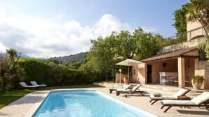 A swimming pool with lounge chairs and a patio area, surrounded by greenery and a mountain in the background. - Luxury Provencal villa with heated pool and gym in St Paul (Saint-Paul-De-Vence)