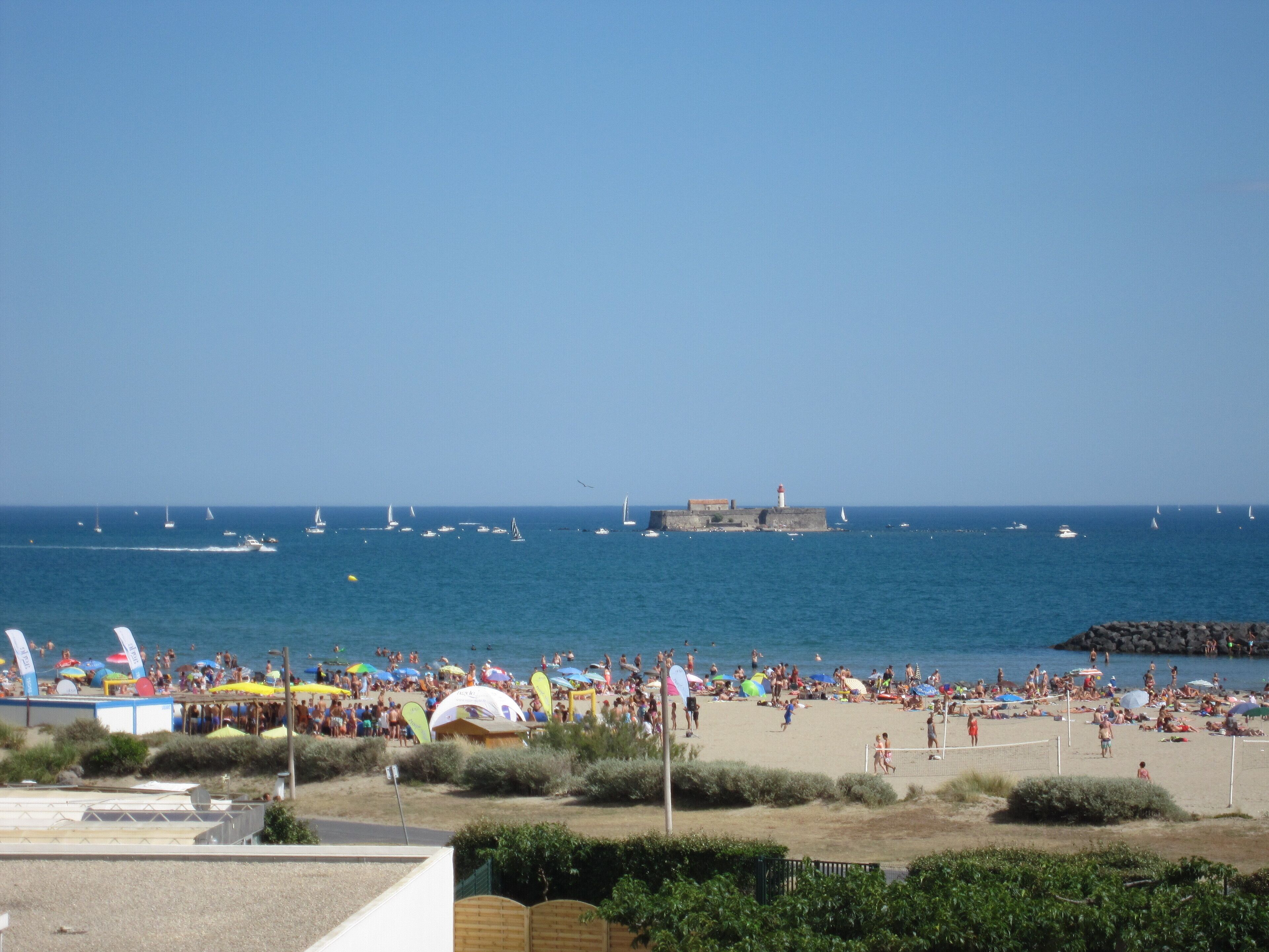 Une plage avec un phare visible au loin, un rivage sablonneux et des bateaux sur l’eau.
