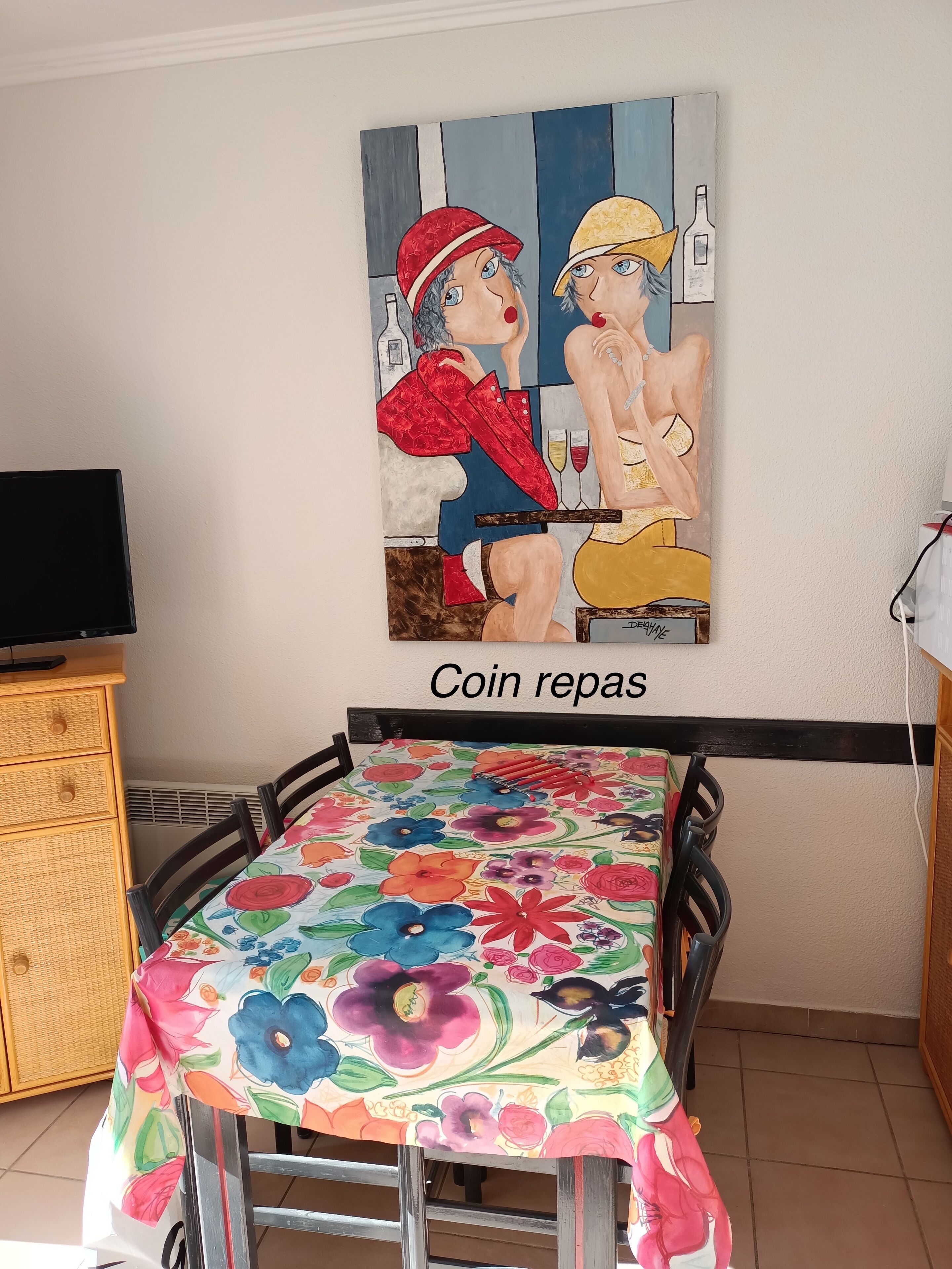 A dining area with a table covered in a floral tablecloth, a painting on the wall, and a sign that reads 'Coin repos.'