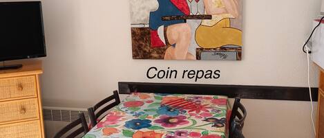 A dining area with a table covered in a floral tablecloth, a painting on the wall, and a sign that reads 'Coin repos.'