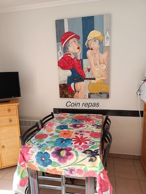 A dining area with a table covered in a floral tablecloth, a painting on the wall, and a sign that reads "Coin repas".