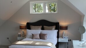 A bedroom with a sloped ceiling, a large bed with multiple pillows, bedside tables with lamps, a small desk with a mirror, and a window providing natural light. - Open Plan Holiday Cottage Within Walking Distance Of Westbourne And Beach (Poole)