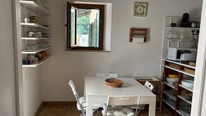 Una cocina compacta con muebles blancos, una mesa de comedor con sillas, una lámpara colgante y una ventana que da a zonas verdes.