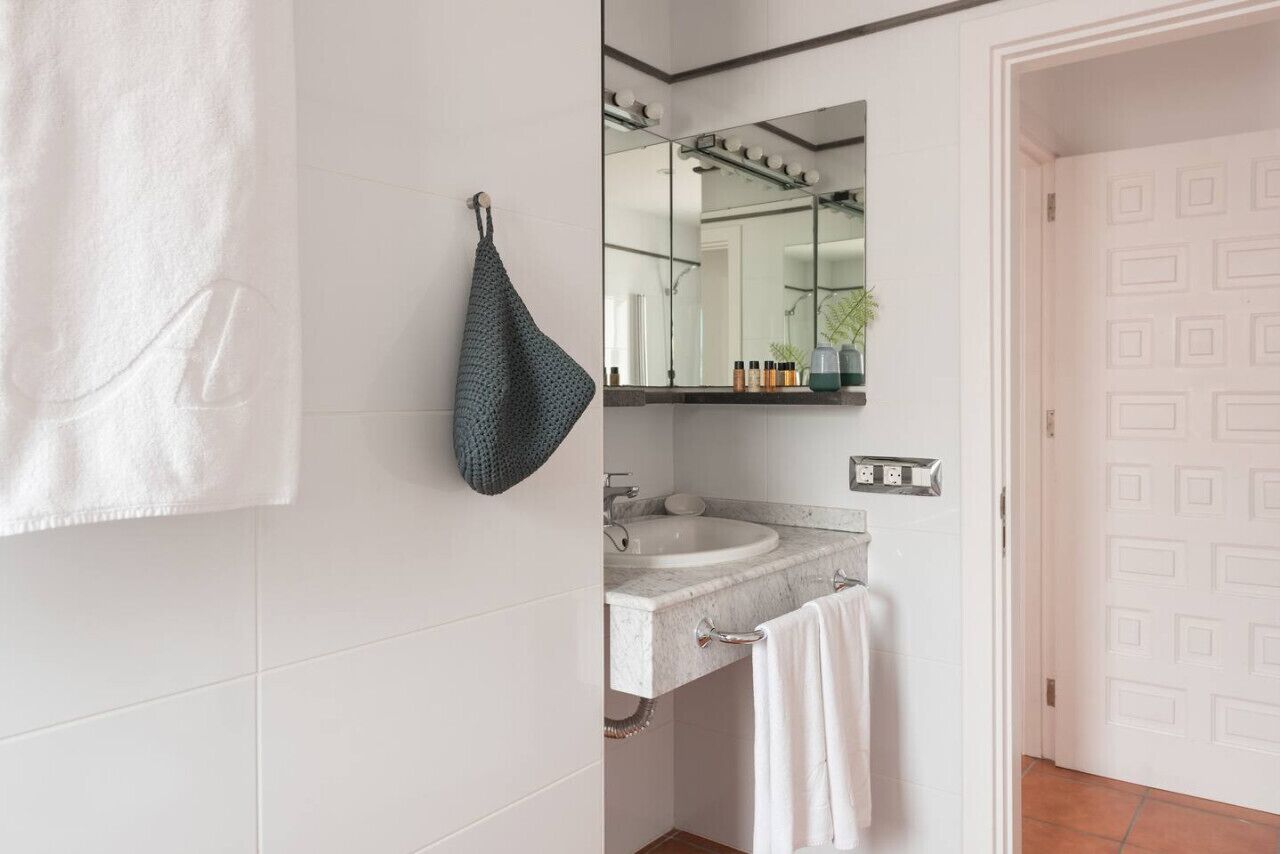 A bathroom with a white tiled wall, a white towel hanging on the wall, a dark towel on a hook, a sink with a mirror above it, and a door with a geometric pattern.