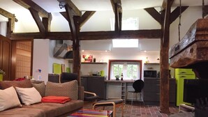 Una sala de estar acogedora con un sofá, una mesa de centro colorida y una chimenea. Hay una zona de cocina con un ventanal y un armario verde.