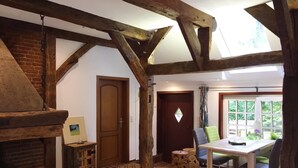 Un interior rústico con vigas de madera a la vista, una chimenea, una zona de comedor con mesa y sillas, y vistas a través de una ventana.