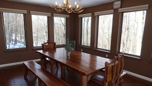 Un comedor con una gran mesa de madera, bancos y sillas. Hay ventanas amplias con vistas a árboles.
